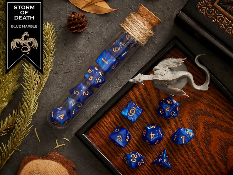 May include: A set of blue and gold dice with a dragon logo, "Storm of Death", and "Blue Marble" text. The dice are in a glass test tube with a cork stopper and are on a wooden surface with a miniature dragon figurine.