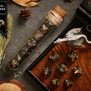 May include: A set of black and gold dice in a glass test tube with a cork stopper. The dice are on a wooden tray with a small dragon figurine. The text "Eternal Darkness Brown + Gold" is on a black label.