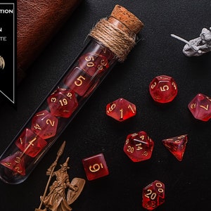 May include: A glass test tube filled with red dice with gold numbers. The test tube is wrapped with twine and has a cork stopper. The dice are scattered around the test tube on a black surface. The text "HEALING POTION BLOOD OF DRAGON RED + WHITE" is on a brown leather label.