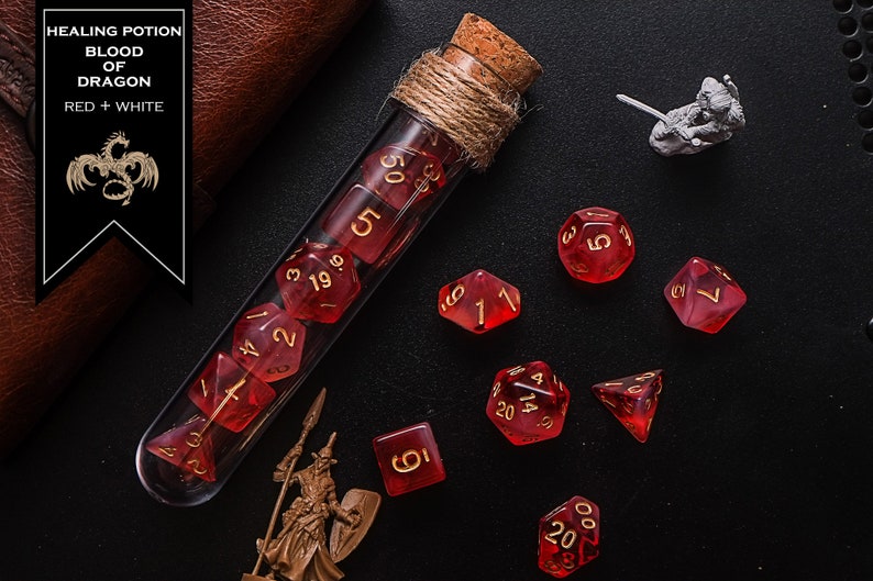 May include: A glass test tube filled with red dice with gold numbers. The test tube is wrapped with twine and has a cork stopper. The dice are scattered around the test tube on a black surface. The text "HEALING POTION BLOOD OF DRAGON RED + WHITE" is on a brown leather label.