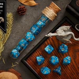 May include: A set of blue and gold dice with a turquoise marble finish. The dice are in a glass tube with a cork stopper and a string tied around it. The dice are on a wooden tray with a small dragon figurine. The text "SPEARS STORM TURQUOISE MARBLE" is on a black label.