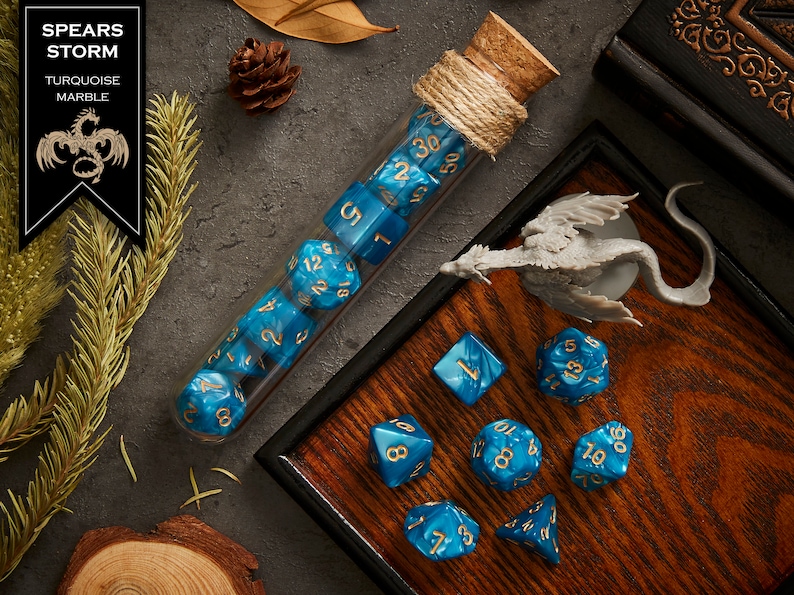 May include: A set of blue and gold dice with a turquoise marble finish. The dice are in a glass tube with a cork stopper and a string tied around it. The dice are on a wooden tray with a small dragon figurine. The text "SPEARS STORM TURQUOISE MARBLE" is on a black label.