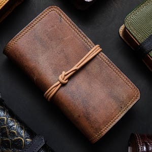 Könnte beinhalten: Ein braunes Lederjournal mit einem beigefarbenen Lederband, das um ihn herumgebunden ist. Das Journal hat einen abgenutzten Look und ist an den Rändern vernäht.