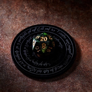 May include: A green twenty-sided die with gold numbers sits on a black circular coaster with an inscription in a circular pattern. The coaster is on a brown surface.