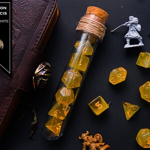 May include: A glass test tube filled with yellow and white dice, with a cork stopper and twine. The dice are numbered 1-6 and 18-20. The test tube is on a black surface with other dice scattered around it. The text "LUCK POTION FELIX FELICIS YELLOW + WHITE" is visible in the top left corner of the image.