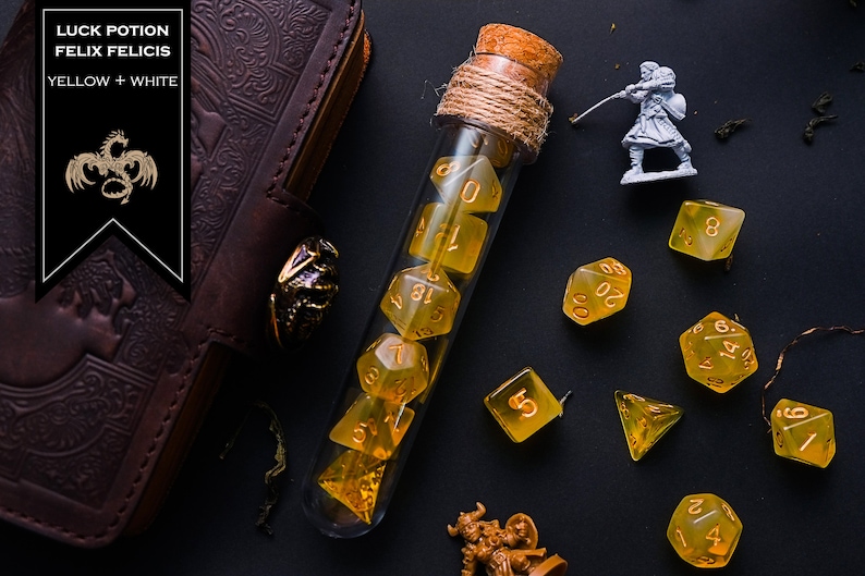 May include: A glass test tube filled with yellow and white dice, with a cork stopper and twine. The dice are numbered 1-6 and 18-20. The test tube is on a black surface with other dice scattered around it. The text "LUCK POTION FELIX FELICIS YELLOW + WHITE" is visible in the top left corner of the image.