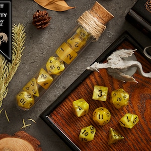 May include: A set of yellow dice with black numbers in a glass test tube with a cork stopper and twine. The dice are on a wooden tray with a small gray dragon figurine. The text "LIGHT OF DIVINITY TUSCANY MARBLE" is on a black label.