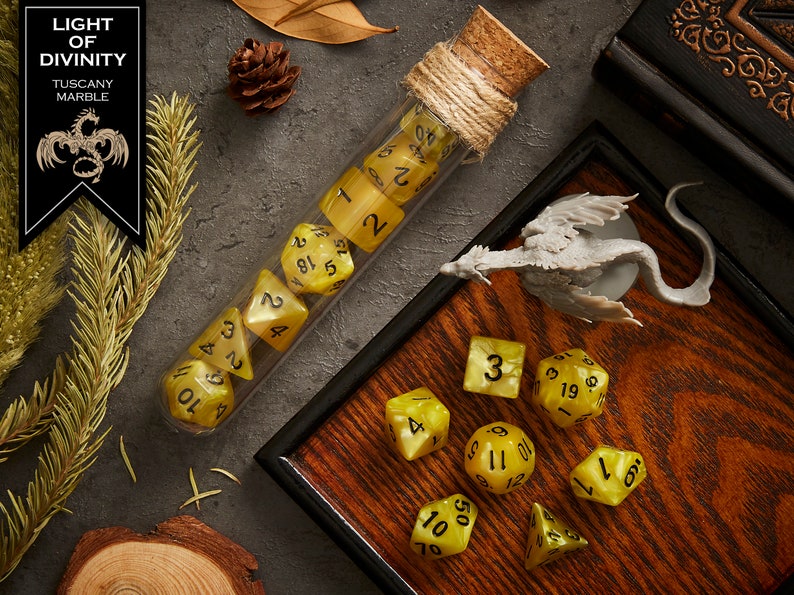 May include: A set of yellow dice with black numbers in a glass test tube with a cork stopper and twine. The dice are on a wooden tray with a small gray dragon figurine. The text "LIGHT OF DIVINITY TUSCANY MARBLE" is on a black label.