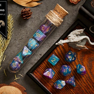 May include: A set of blue and purple dice with gold numbers in a glass tube with a cork stopper. The dice are on a wooden tray with a small dragon figurine. The text "RAGE OF DRAGONS BLUE + PURPLE" is on a black banner.