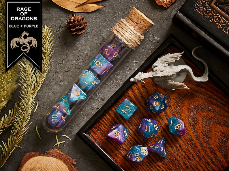May include: A set of blue and purple dice with gold numbers in a glass tube with a cork stopper. The dice are on a wooden tray with a small dragon figurine. The text "RAGE OF DRAGONS BLUE + PURPLE" is on a black banner.