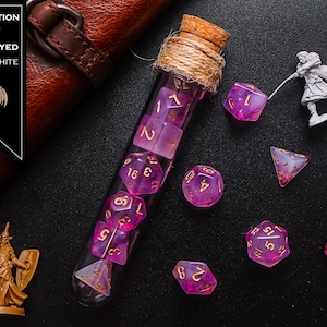 May include: A glass test tube filled with purple and white dice with gold numbers. The test tube is corked and tied with twine. The dice are scattered around the test tube on a black surface. The text "HYBRID POTION BLOOD OF THE BETRAYED PURPLE + WHITE" is on a black label.
