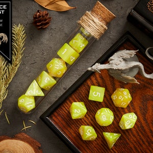 May include: A set of yellow marble dice with white numbers, some in a glass test tube with a cork stopper and twine, and others on a wooden tray with a gray dragon figurine. The text "JUSTICE BLAST YELLOW MARBLE" is on a black label.