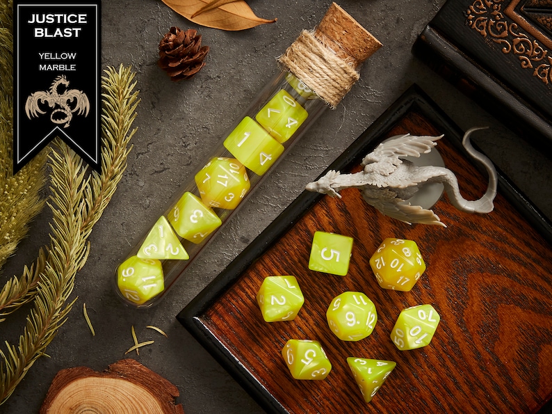 May include: A set of yellow marble dice with white numbers, some in a glass test tube with a cork stopper and twine, and others on a wooden tray with a gray dragon figurine. The text "JUSTICE BLAST YELLOW MARBLE" is on a black label.