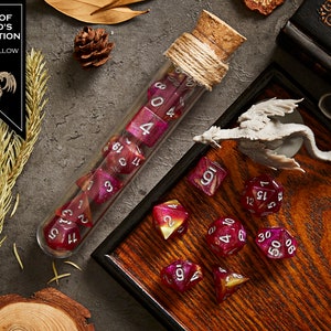 May include: A set of pink and yellow dice with silver numbers, in a glass test tube with a cork stopper, on a wooden tray with a small dragon figurine. The dice are part of a set called "Lord of Blood's Exultation".
