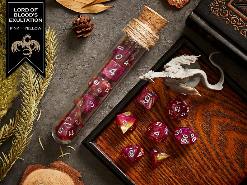 May include: A set of pink and yellow dice with silver numbers, in a glass test tube with a cork stopper, on a wooden tray with a small dragon figurine. The dice are part of a set called "Lord of Blood's Exultation".