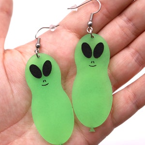 May include: A pair of green alien-shaped earrings with black eyes and a smiling mouth. The earrings are made of a translucent material and hang from silver-colored ear wires. The alien design resembles a balloon.