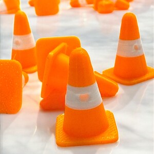 May include: An assortment of small, bright orange traffic cones with white reflective bands. The cones are scattered across a reflective surface, some upright and others lying down. The cones have a square base.