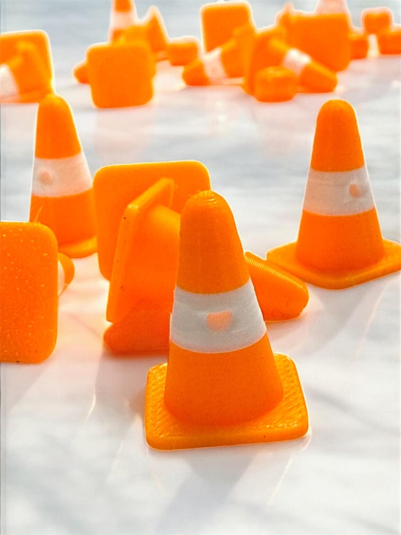 May include: An assortment of small, bright orange traffic cones with white reflective bands. The cones are scattered across a reflective surface, some upright and others lying down. The cones have a square base.