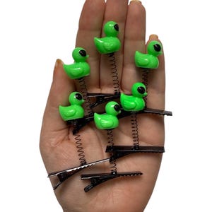 May include: A collection of six green duck hair clips with black eyes and beaks. Each duck is attached to a black metal clip with a silver spring. The clips are arranged in a fan shape.
