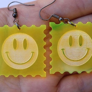 May include: A pair of yellow smiley face earrings with a green and white swirl pattern on one earring. The earrings are made of acrylic and have a simple hook closure.