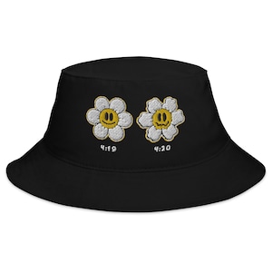 Puede incluir: Sombrero de pescador negro con dos flores blancas bordadas. Las flores tienen caras sonrientes amarillas. El texto "4:19" y "4:20" está bordado debajo de las flores.