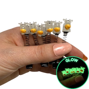 May include: A set of six cow-shaped novelty items with yellow noses and black springs. The items are held in a hand. A glowing inset shows the items glowing green in the dark. The text "ONE STOP RAVE" is in the upper right corner.