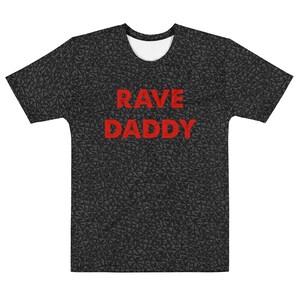 以下が含まれることがあります： 暗いグレーのテクスチャパターンが施された、黒の半袖Tシャツ。Tシャツの前面には赤い文字で「RAVE DADDY」とプリントされています。
