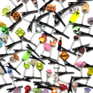 Pu&ograve; includere: Una collezione di fermagli per capelli neri con vari ciondoli colorati. I ciondoli includono alieni, funghi, anatre e segnali stradali. Alcuni ciondoli presentano il testo "HOT" e "BUMP AHEAD".