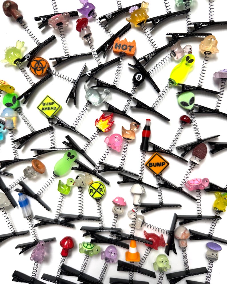 Pu&ograve; includere: Una collezione di fermagli per capelli neri con vari ciondoli colorati. I ciondoli includono alieni, funghi, anatre e segnali stradali. Alcuni ciondoli presentano il testo "HOT" e "BUMP AHEAD".
