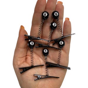 Puede incluir: Pinzas para el pelo negras con un resorte y un diseño de bola ocho negra. Las pinzas se sostienen en una mano con esmalte de uñas naranja. La bola ocho es negra con un número ocho blanco.