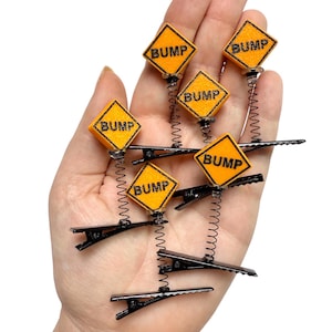 May include: Novelty hair clips with orange diamond signs reading "BUMP" in black. Each clip has a black base and a silver spring, like miniature road signs. The clips are designed to be fun and eye-catching.