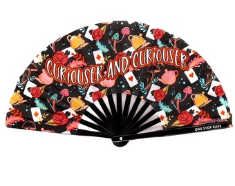 May include: A black folding fan with a white and black handle. The fan is decorated with a black background and a colorful pattern of teacups, roses, playing cards, and mushrooms. The text "Curiouser and Curiouser" is printed in orange on the fan.