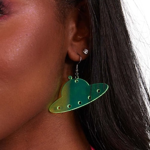 May include: Green and yellow iridescent UFO-shaped earrings. The earrings have a circular design with multiple holes and a small antenna. The earrings are hanging from a silver hook. The earrings are a fun and unique accessory.