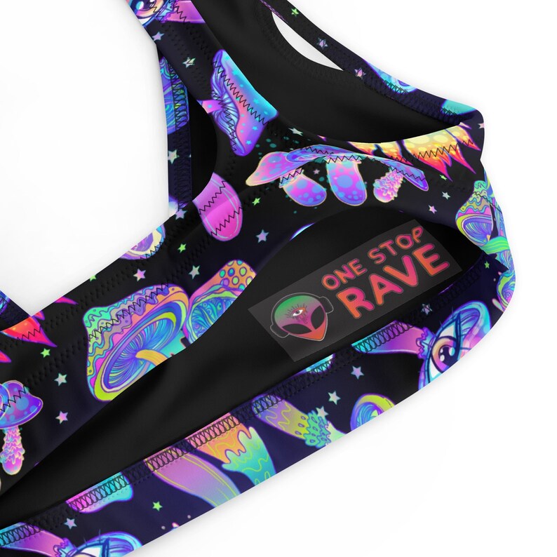 May include: Black bikini top with a vibrant, psychedelic mushroom and star pattern. The design features neon colors and the text "ONE STOP RAVE" on a label. The top is made of a smooth, likely synthetic fabric.