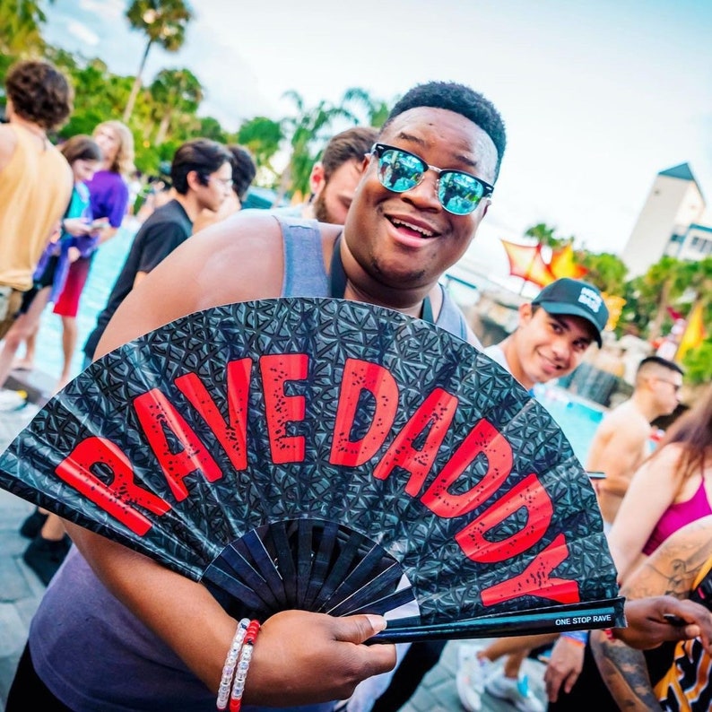 K&ouml;nnte beinhalten: Eine l&auml;chelnde Person h&auml;lt einen schwarz-grauen F&auml;cher mit den roten Worten "RAVE DADDY". Der F&auml;cher hat ein geometrisches Muster. Die Person tr&auml;gt eine Sonnenbrille und ein graues Tanktop. Andere Personen sind im Hintergrund.