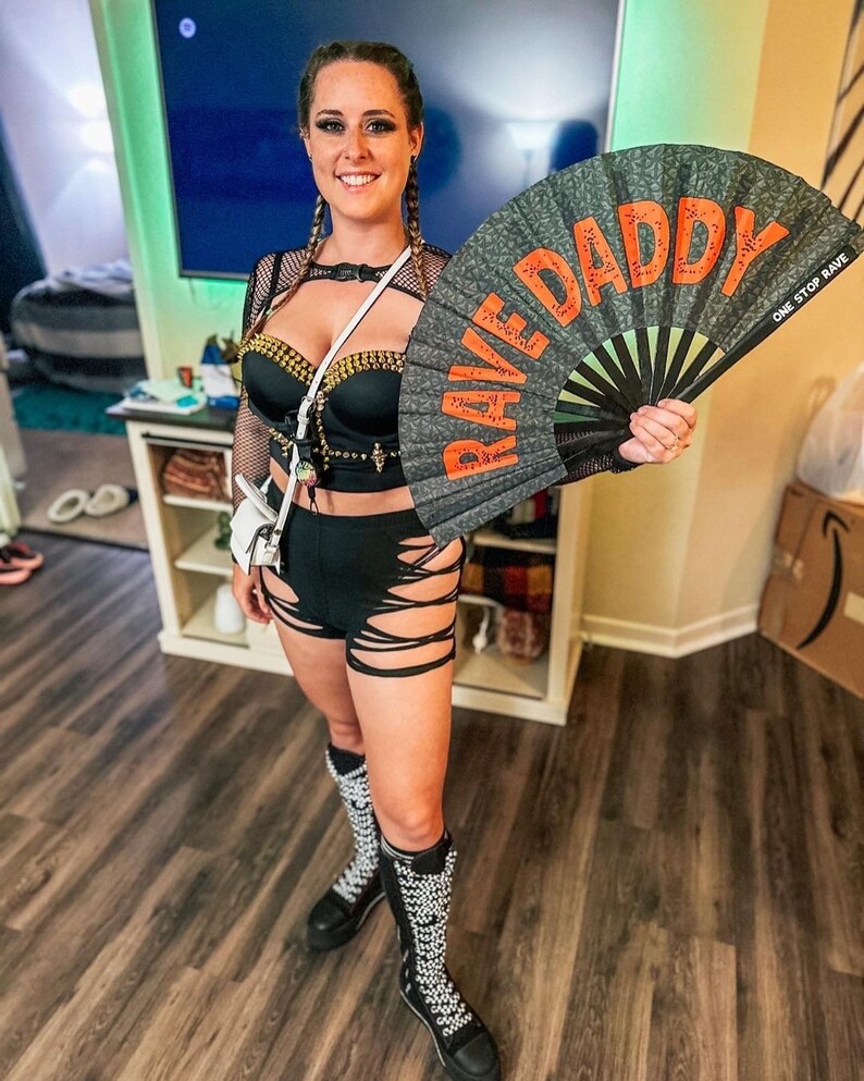K&ouml;nnte beinhalten: Eine Person in einem schwarzen Outfit, die einen F&auml;cher mit den Worten "RAVE DADDY" in Orange h&auml;lt. Das Outfit umfasst ein mit Nieten besetztes Oberteil, Cut-Out-Shorts und High-Top-Stiefel. Der F&auml;cher tr&auml;gt auch den Text "ONE STOP RAVE".