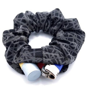 May include: A dark gray scrunchie with a geometric pattern. The scrunchie has a hidden pocket containing a small, silver tube and two lighters, one red and one blue.