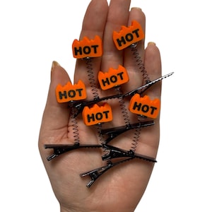 May include: A collection of black hair clips with orange, flame-shaped accents. Each clip features the word "HOT" in black lettering. The clips are attached to a coiled spring.