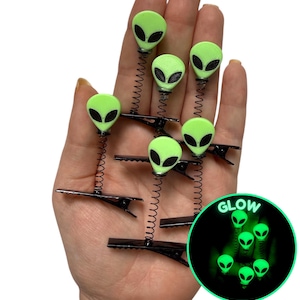 May include: A collection of neon green alien head hair clips with black springs and clips. The alien heads have black eyes and are displayed on a hand. A circular inset shows the alien heads glowing green in the dark. The text "ONE STOP RAVE" is in the top right corner.