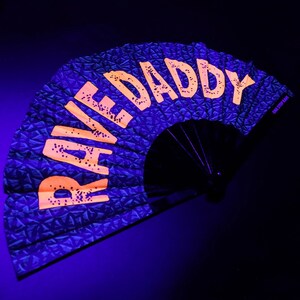 K&ouml;nnte beinhalten: Ein schwarzer Faltf&auml;cher mit den Worten "RAVE DADDY" in leuchtend orangefarbenen Blockbuchstaben. Der Stoff des F&auml;chers hat ein strukturiertes, geometrisches Muster in Dunkelblau. Der F&auml;cher ist ge&ouml;ffnet und zeigt seine volle Form vor dunklem Hintergrund.
