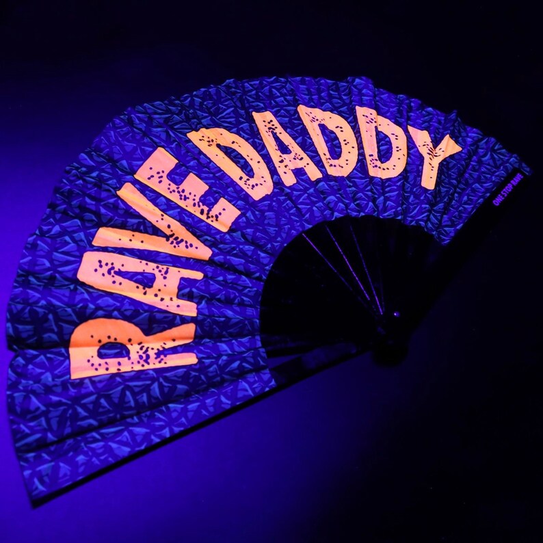 K&ouml;nnte beinhalten: Ein schwarzer Faltf&auml;cher mit den Worten "RAVE DADDY" in leuchtend orangefarbenen Blockbuchstaben. Der Stoff des F&auml;chers hat ein strukturiertes, geometrisches Muster in Dunkelblau. Der F&auml;cher ist ge&ouml;ffnet und zeigt seine volle Form vor dunklem Hintergrund.