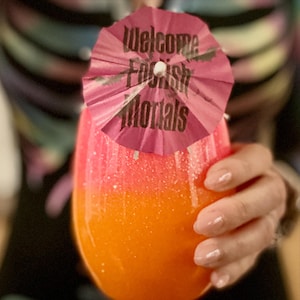 May include: A vibrant, layered cocktail in a tulip-shaped glass, transitioning from orange to pink. A pink paper umbrella with the text "Welcome Foolish Mortals" adorns the drink. The drink is held by a person.