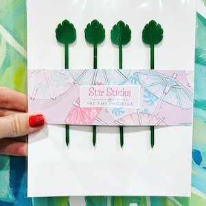 May include: Four green plastic stir sticks with a leaf design. The sticks are packaged in a clear plastic bag with a white label that says "Stir Sticks" and "The Tiny Umbrella".