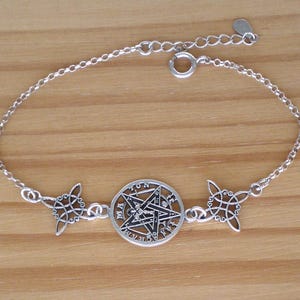 May include: Silver-toned bracelet featuring a central pentagram pendant within a circular frame, flanked by two decorative Celtic knot charms. The adjustable chain is connected with a lobster clasp. The bracelet is displayed on a light brown wooden surface.