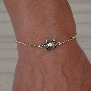 Puede incluir: Un brazalete de cadena de plata con un pequeño dije de tortuga. El brazalete está en la muñeca de una persona.