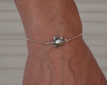 May include: A silver chain bracelet with a small turtle charm. The bracelet is on a person's wrist.