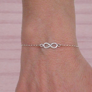 Puede incluir: Un brazalete de plata con un dije de símbolo de infinito.