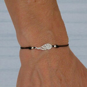 Könnte beinhalten: Ein Armband mit einem silbernen Flügelanhänger und einem schwarzen Band. Das Armband hat ein filigranes Flügeldesign und kleine silberne Perlen. Das Armband wird am Handgelenk getragen.