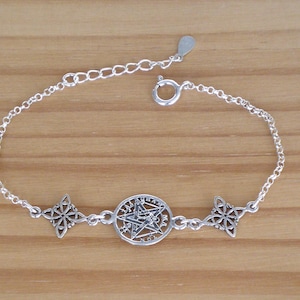 Op de afbeelding: Een zilverkleurige armband met een centrale pentagramhanger, geflankeerd door twee decoratieve sterrenburst bedels. De armband heeft een fijne ketting en een karabijnsluiting. De sieraden worden gepresenteerd op een lichtbruin houten oppervlak.