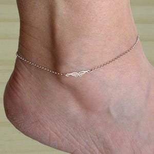 Könnte beinhalten: Ein zartes silbernes Fußkettchen mit einem kleinen Anhänger in Form eines Blattes. Das Fußkettchen besteht aus einer dünnen Kette und ist perfekt zum Schichten mit anderem Schmuck.